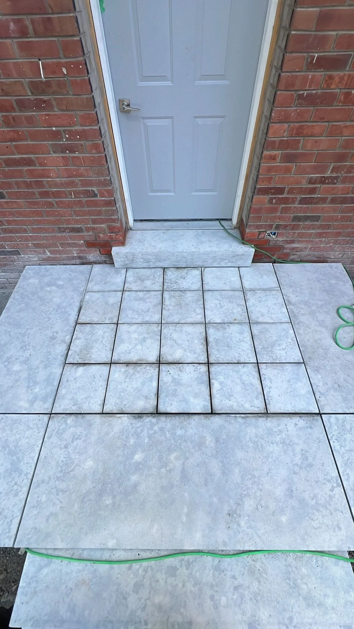 Front porch with a concrete and tiled steps leading to a white door, brick exterior walls, and green extension cords.