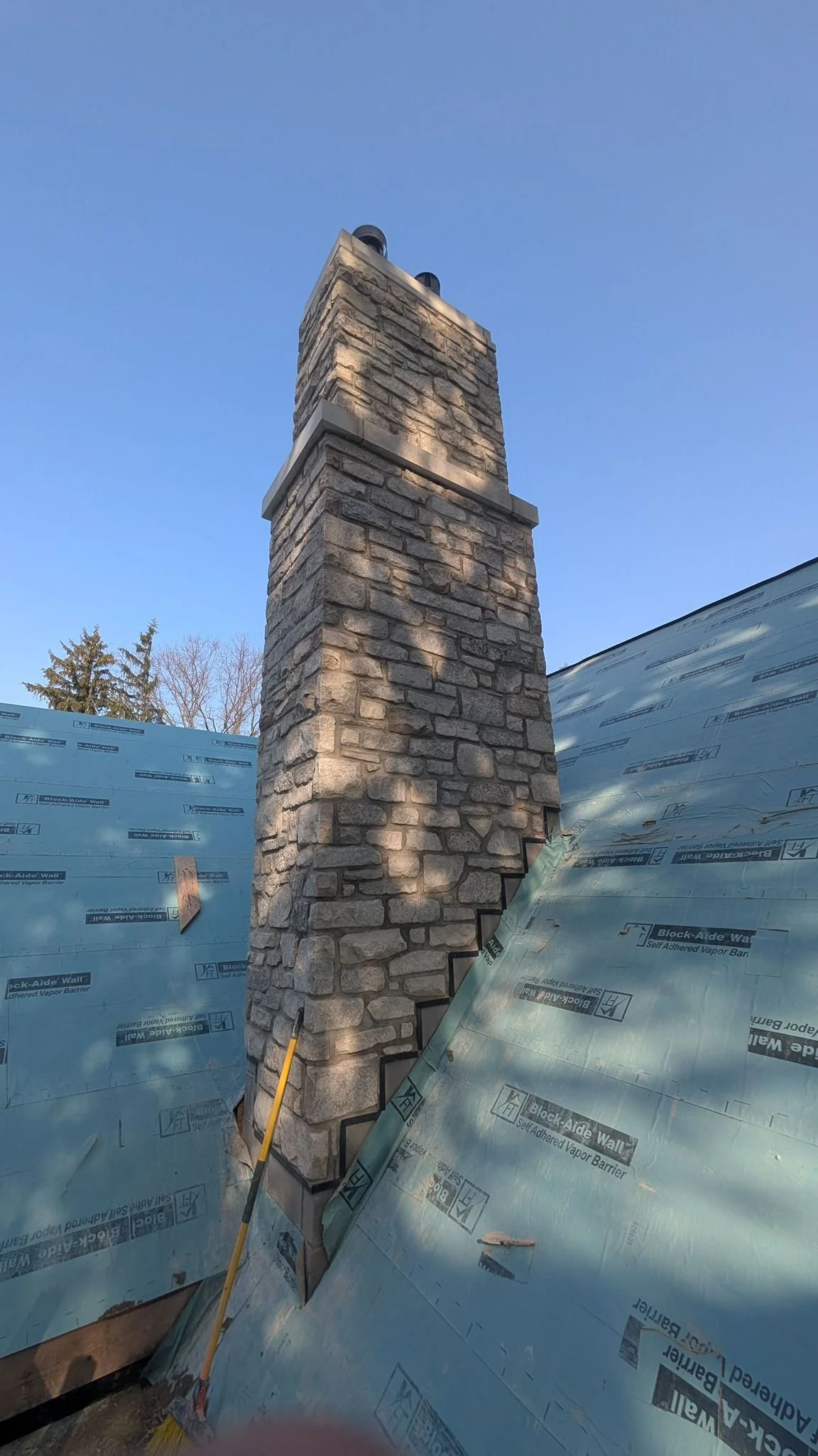 A brick chimney on a house under construction, with blue weather-resistant barrier on the roof and a clear blue sky in the background.