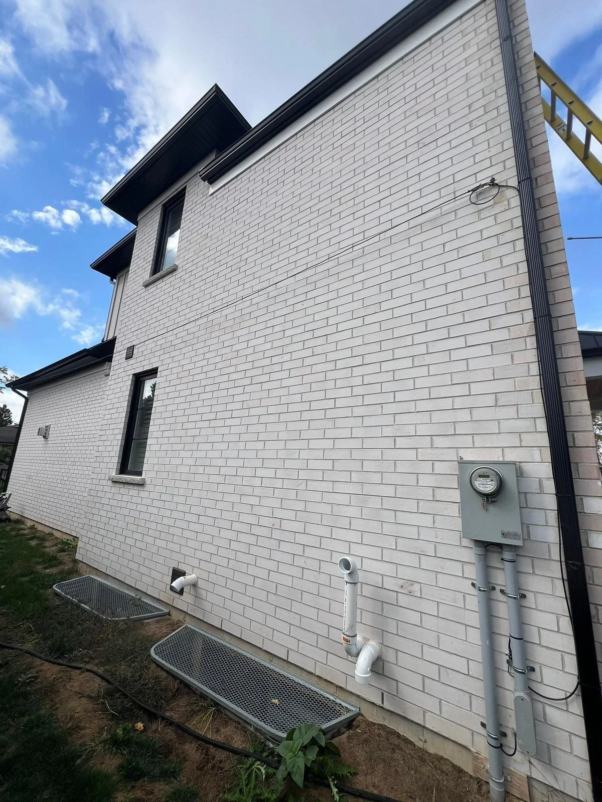 Side of a white brick house with three windows, black trim, and visible utility meters and pipes along the foundation, under a partly cloudy sky.