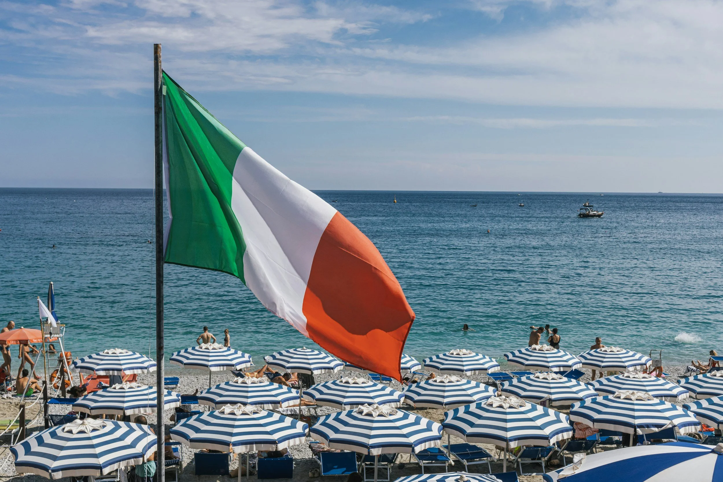 Italian Flag flying over a beach with blue and white striped umbrellas on a sunny day overlooking the Adriatic Sea.