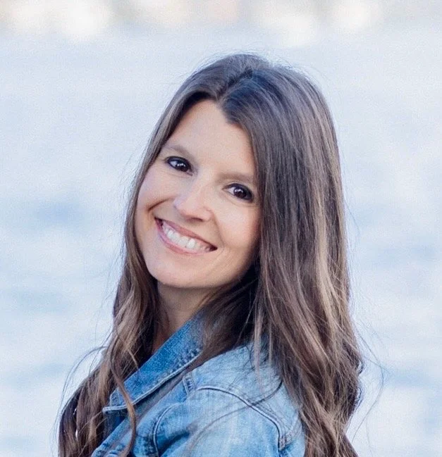 A young woman with long brown hair smiling at the camera with a body of water in the background.
