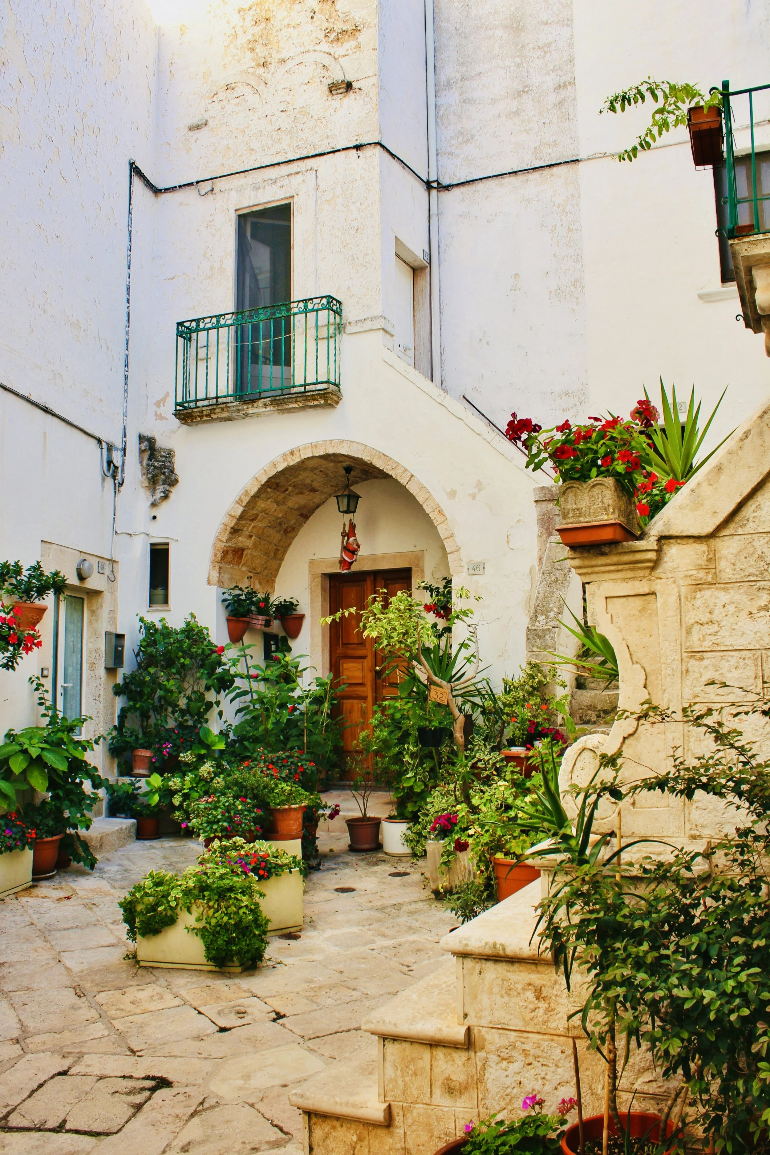 A charming outdoor courtyard of a Mediterranean style house with white stucco walls, featuring potted plants and flowers, a stone archway over the front door, and a small balcony with a green metal railing.