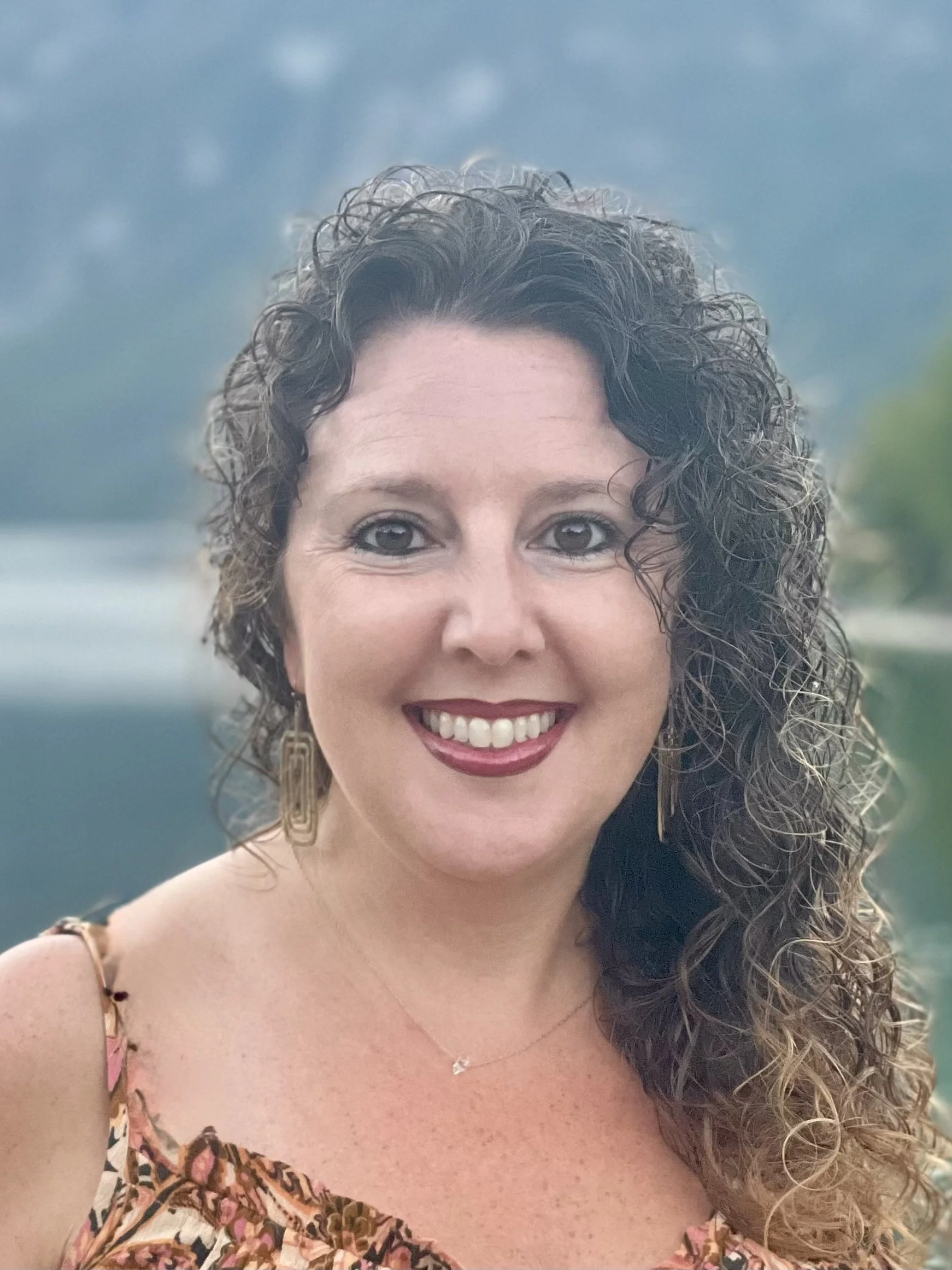 Portrait of a young woman with long, curly brown hair, wearing a black off-the-shoulder top, standing outdoors on a bright day, with a body of water in the background.