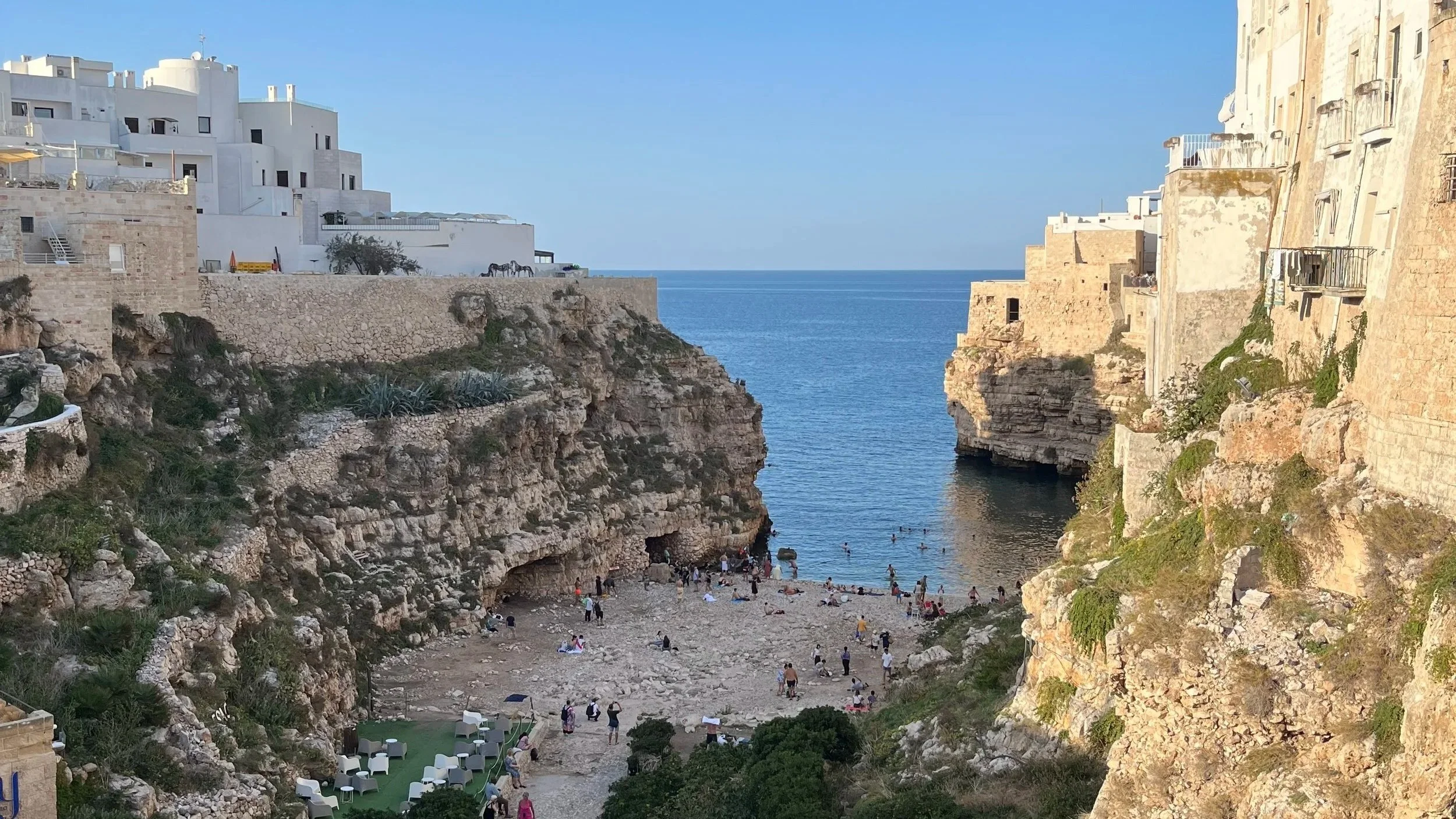 A summer day in Polignano a Mare, Italy. Tourist boats float in the blue sea near crowded beaches and the historic stone bridge.