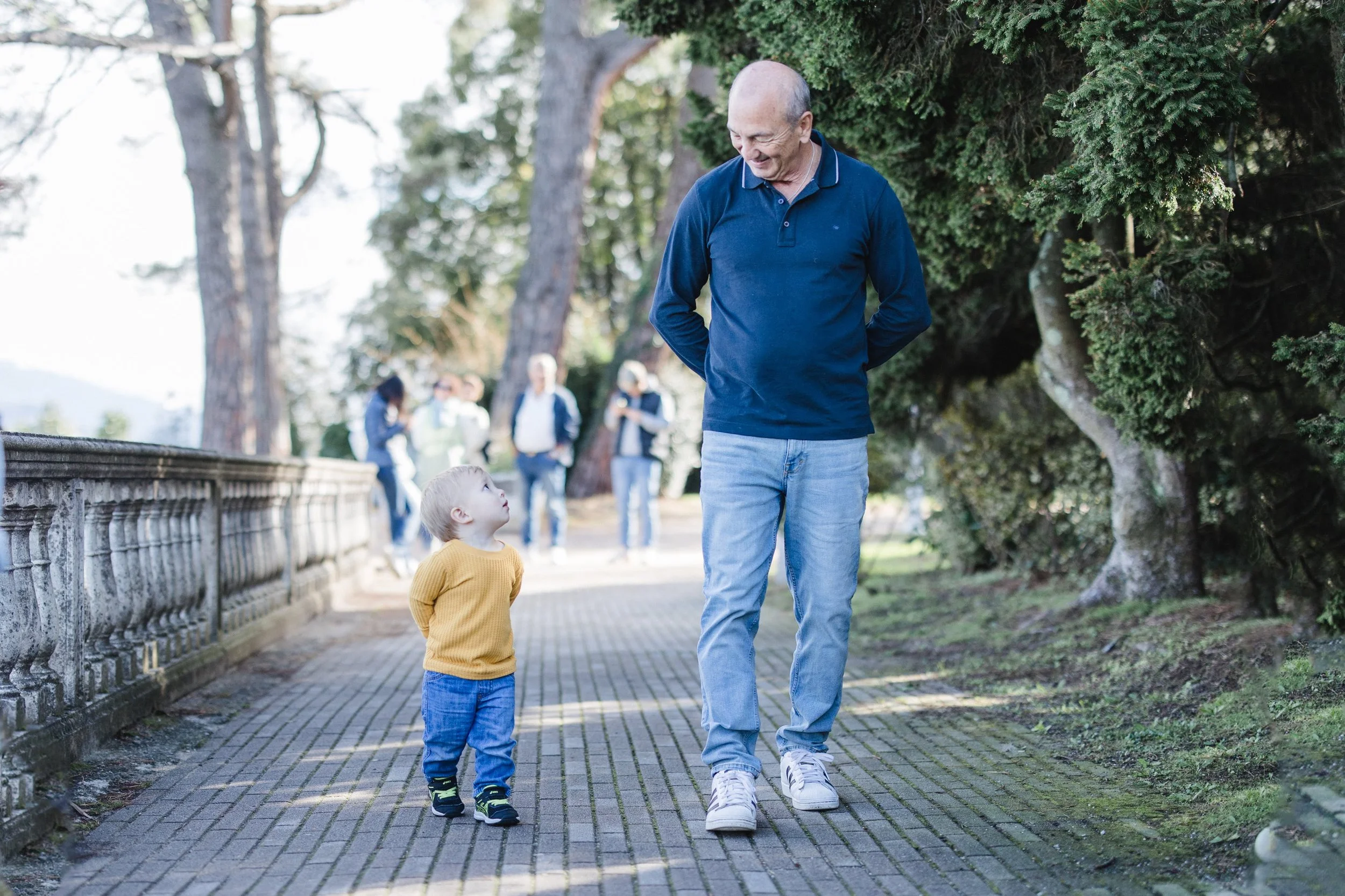 A toddler and grandfather, with their hands behind their backs, taking a stroll down the cobblestone roads in Italy, while a group is onlooking in the background.