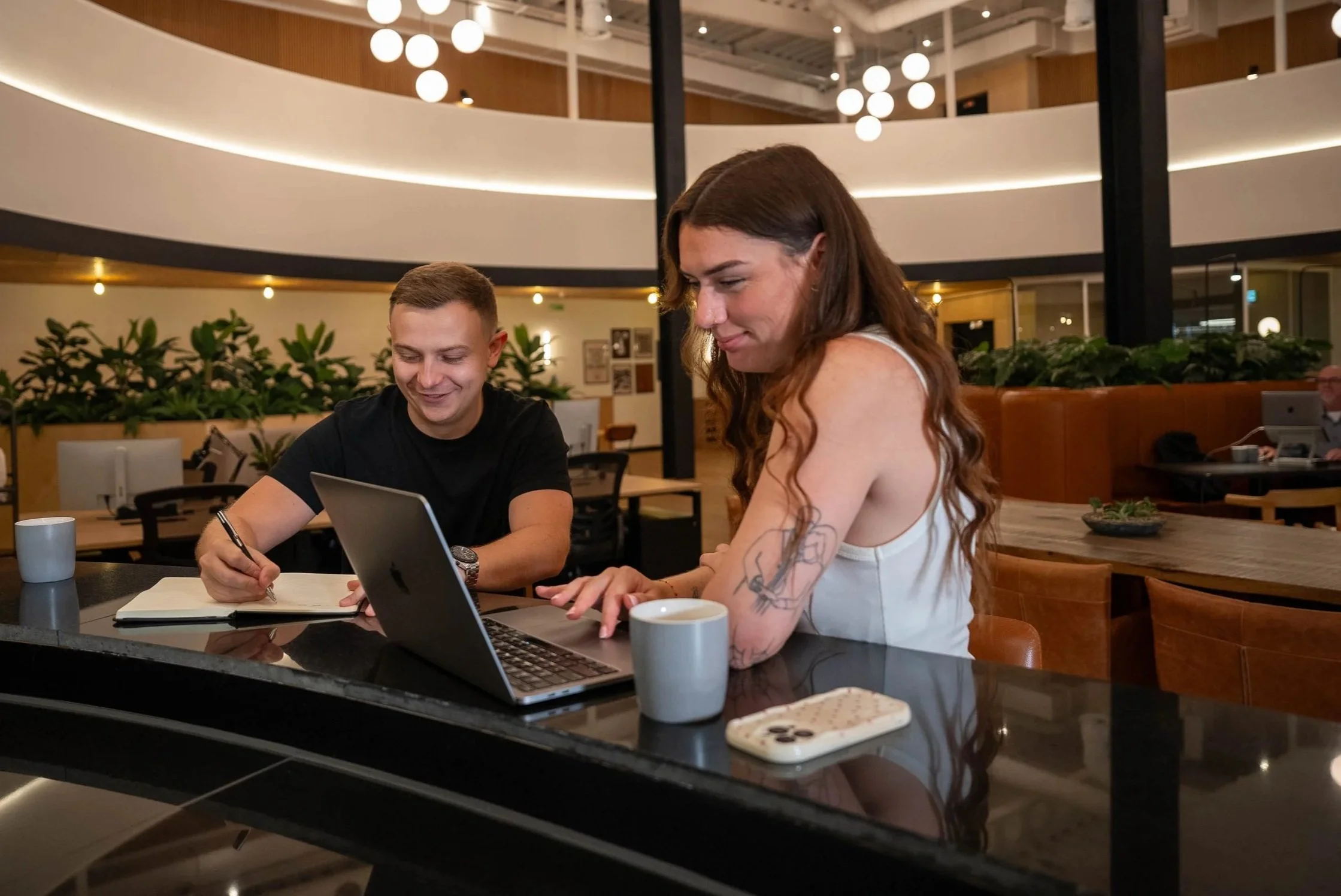 Two people sitting at a table in a modern cafe or restaurant, working together on a laptop. The man is smiling and writing in a notebook, while the woman is typing on the laptop. There are cups, a phone, and a plant on the table. The background features a circular railing, hanging lights, and other patrons.