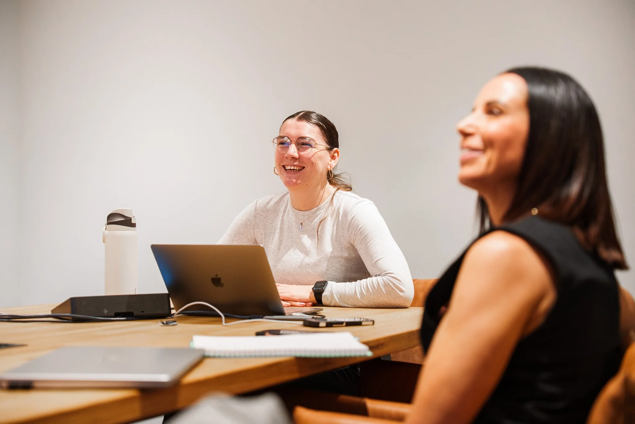 Two women sitting at a wooden table in a meeting room, smiling, with a laptop, notebooks, a water bottle, and a smartphone on the table.