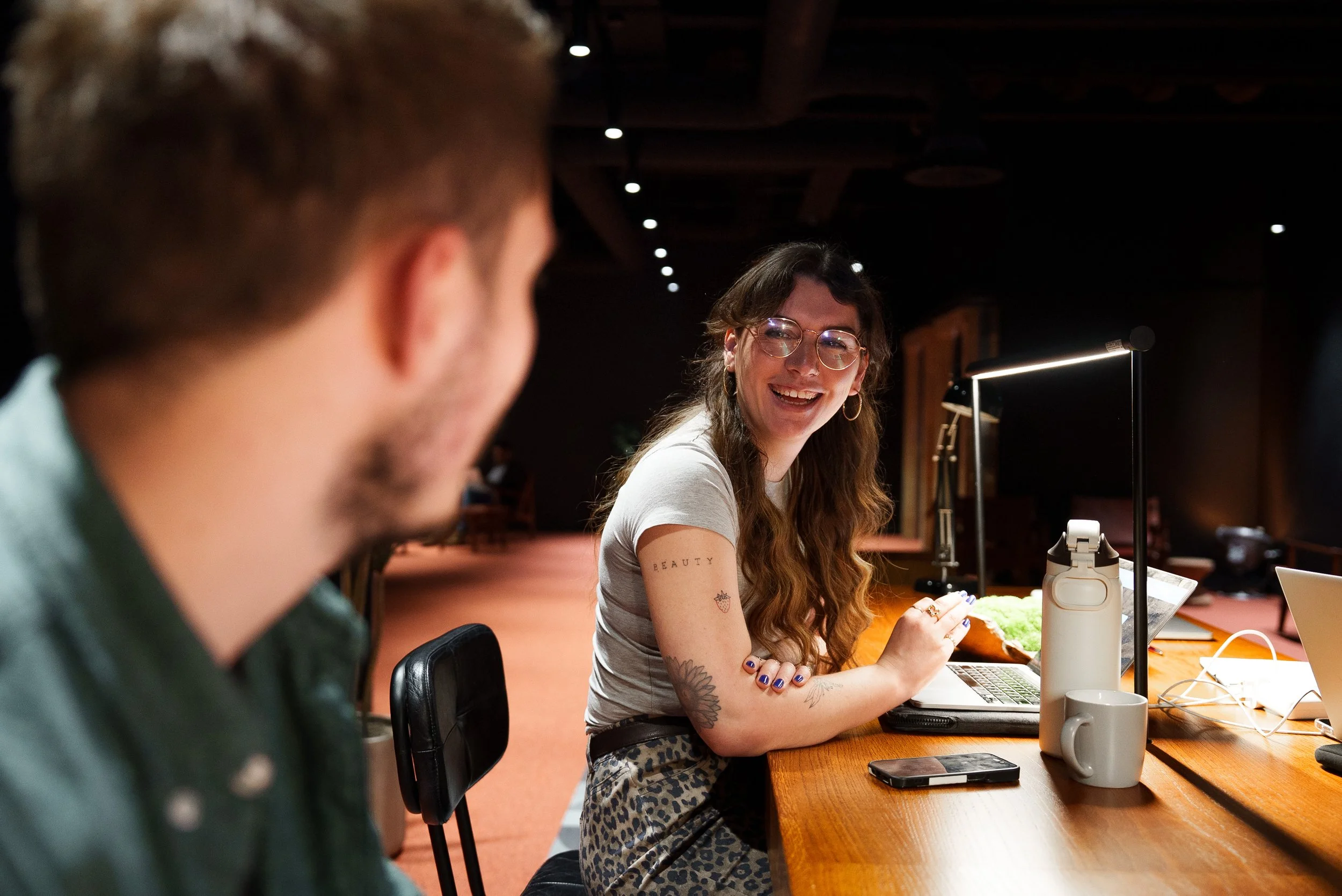 Lily Deal with long wavy hair and glasses, smiling while working on a laptop at a wooden table, with a man blurred in the foreground, in a cozy indoor space.