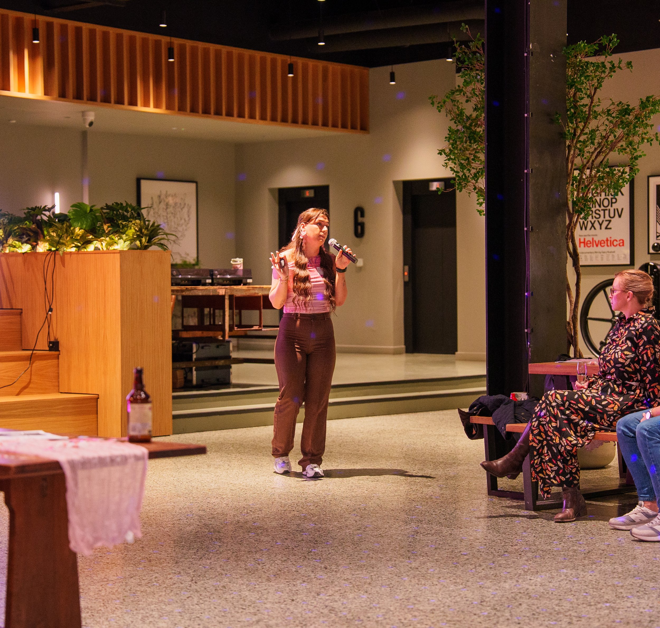 Lily Deal with long wavy hair singing into a microphone on a stage in an indoor venue, with a woman seated on a bench watching. The venue has warm lighting, plants, framed wall art, and a music poster on the wall.