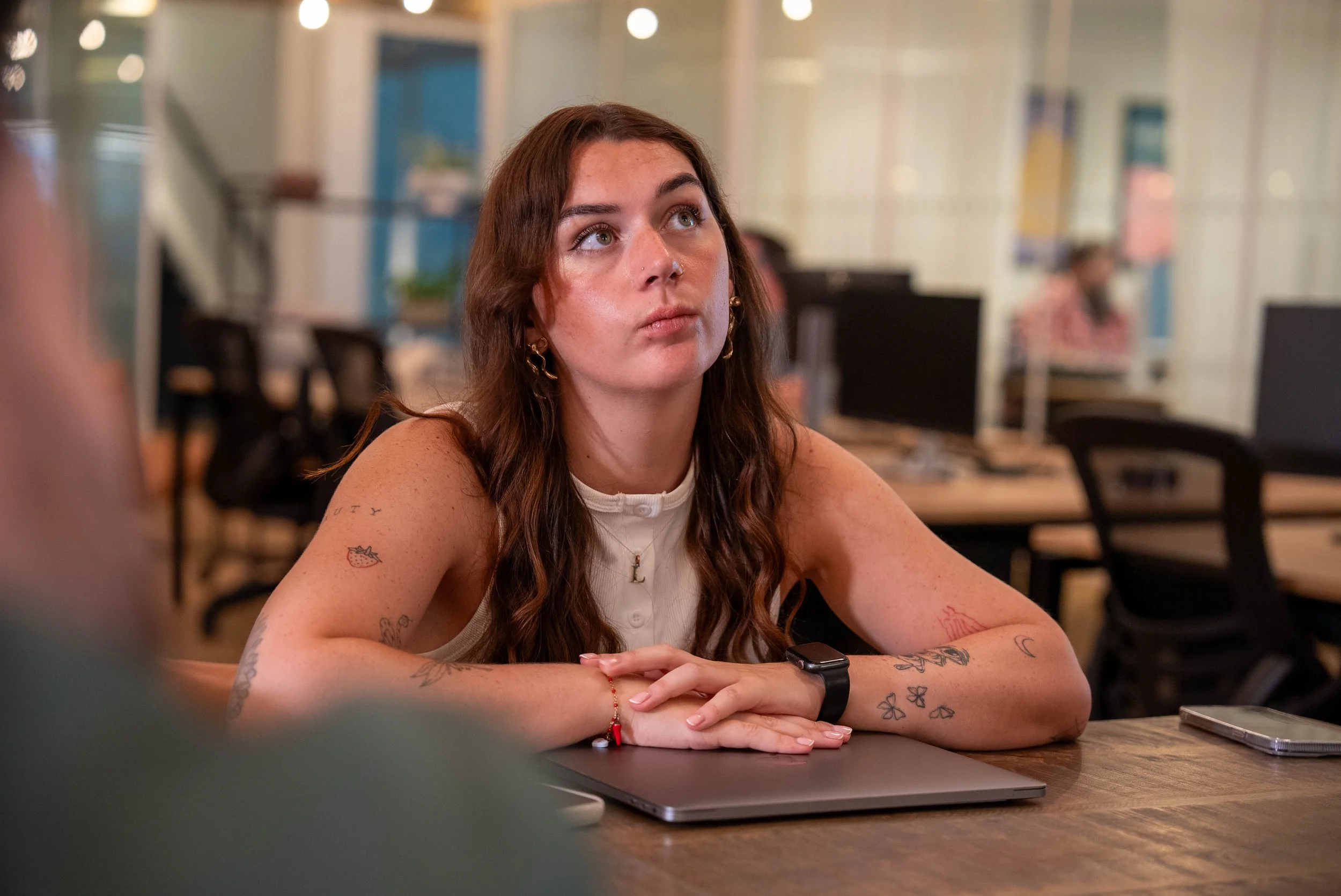 A young woman with long brown hair and tattoos on her arms sitting at a desk in an office, looking up thoughtfully with her hands resting on a closed laptop.