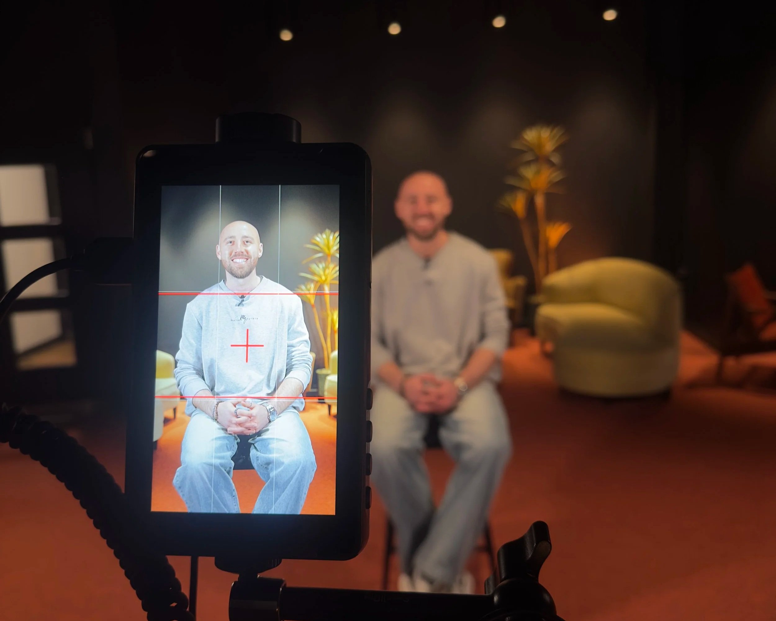 A man with a bald head and beard is sitting on a chair, smiling, in front of a dark background. The photo is being taken with a camera, which shows a clear, focused image of the man.