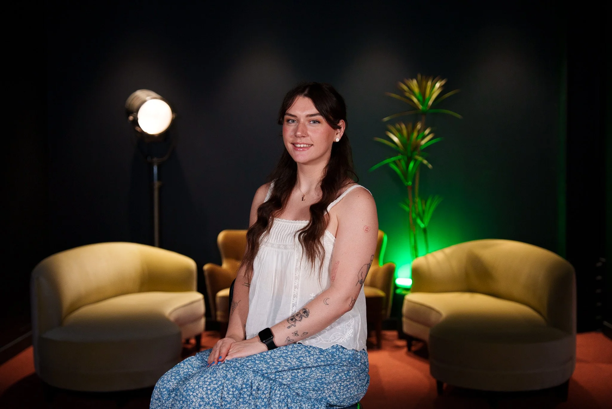 A young woman with long dark hair and tattoos on her arms, wearing a white sleeveless top and blue skirt, sitting in a studio with black walls, yellow curved chairs, and a green-lit plant behind her.