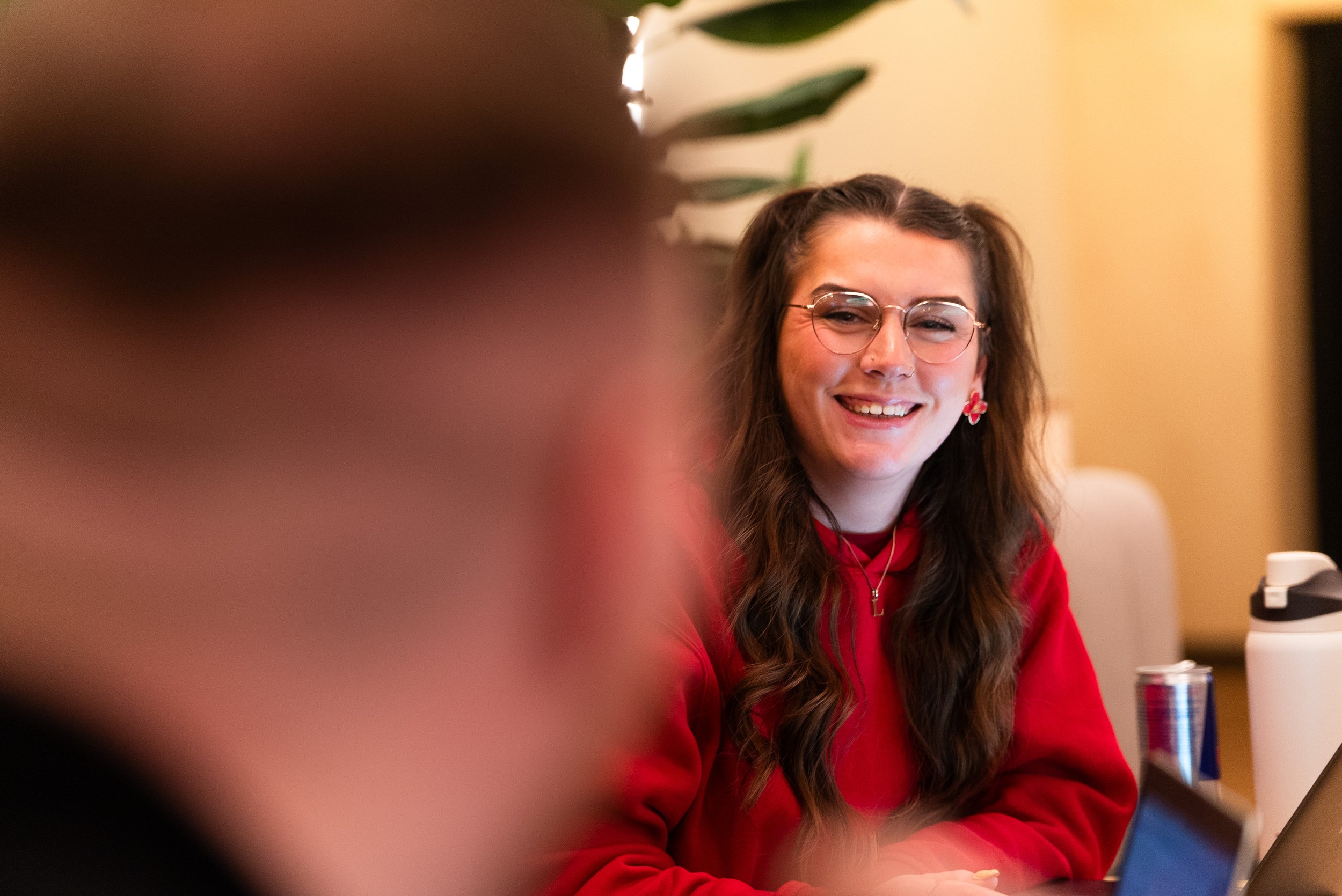 Lily Deal with long wavy brown hair, glasses, and earrings, smiling while sitting at a table, wearing a red hoodie. There is a plant in the background and a laptop, a water bottle, and a can on the table.