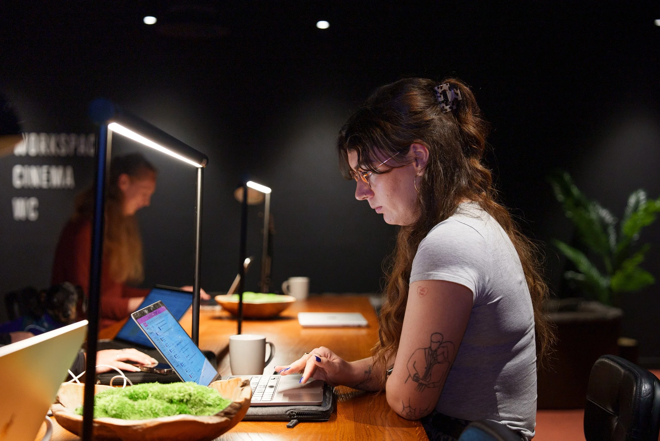 A young woman with tattoos and glasses working on a laptop at a wooden table in a dimly lit room, with other people working on laptops in the background.