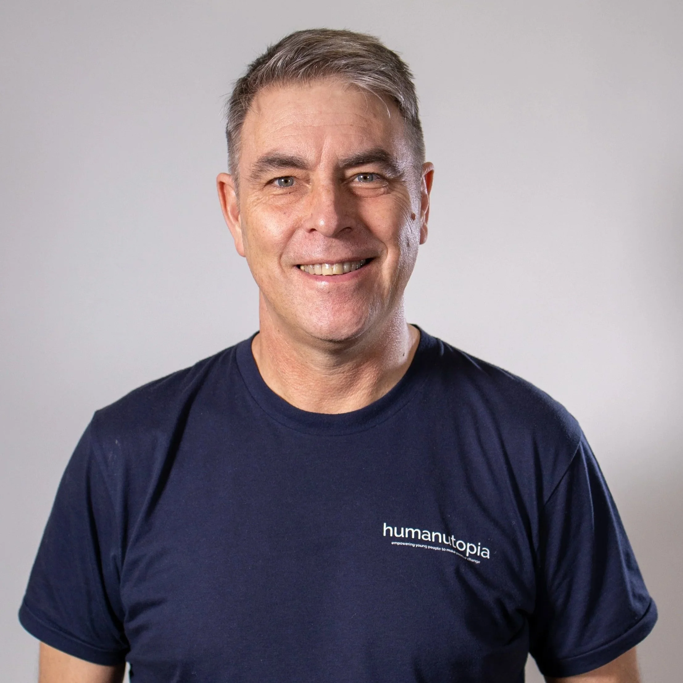 A smiling man with short gray hair, wearing a navy blue T-shirt with the logo 'humanutopia' on it, standing against a plain gray background.