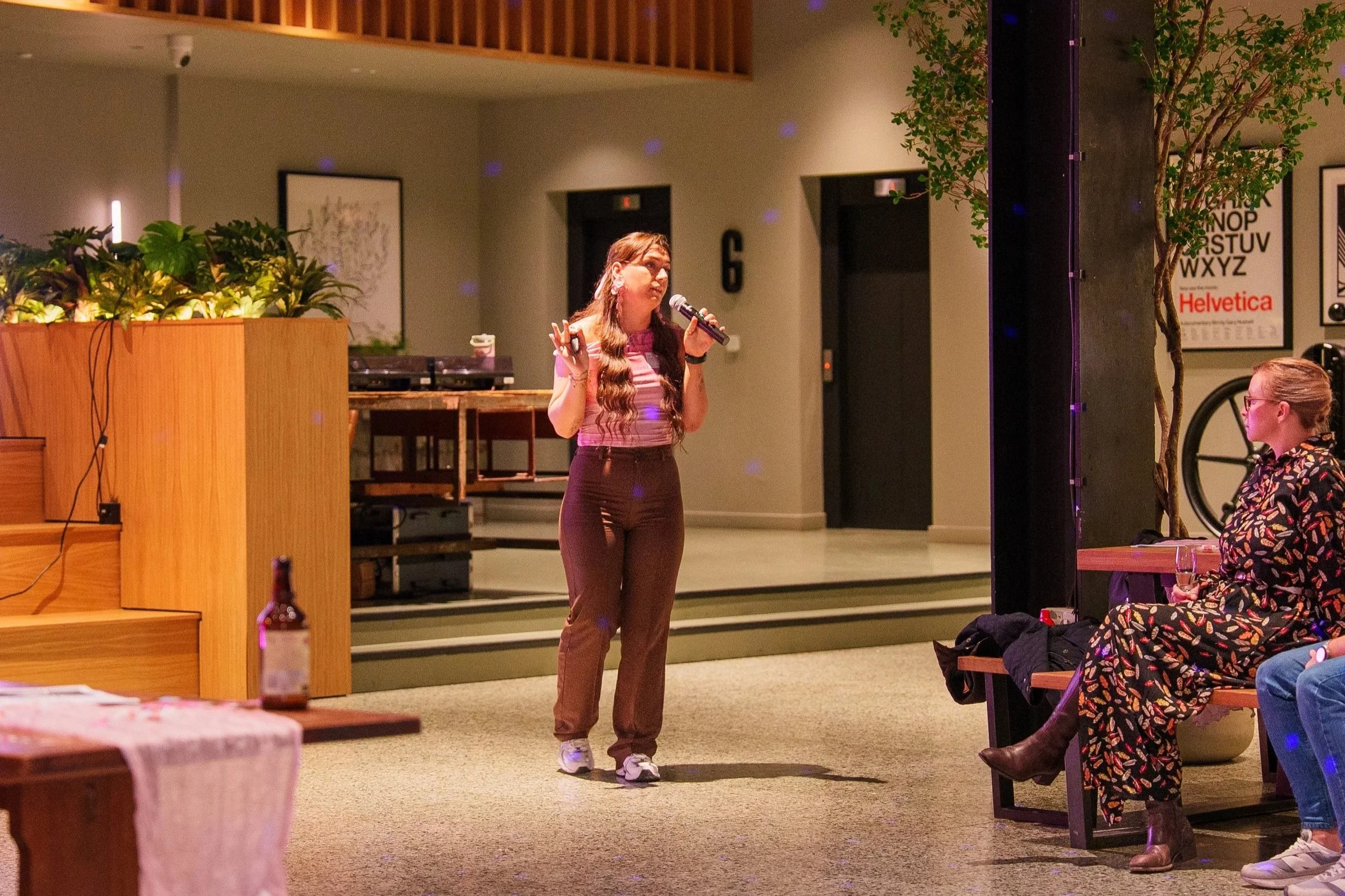 A woman with long wavy hair in a pink striped top and brown pants is speaking into a microphone on a small stage in a casual indoor setting, with a viewer seated nearby.