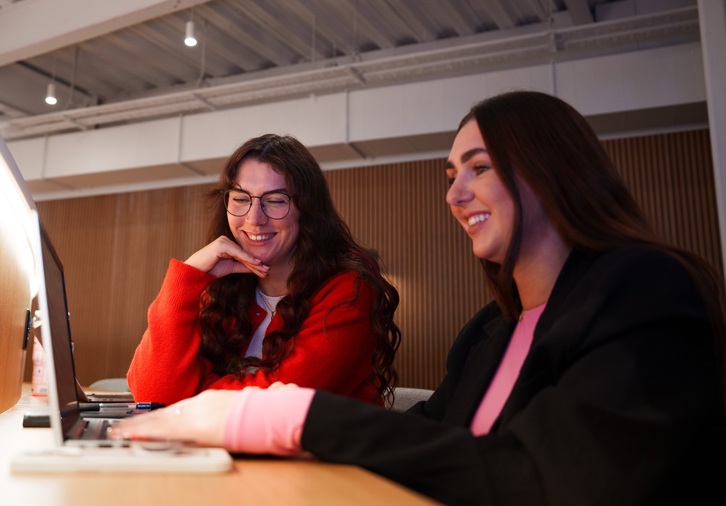 Two women looking at a laptop screen, smiling and engaging in conversation, in a modern indoor setting.