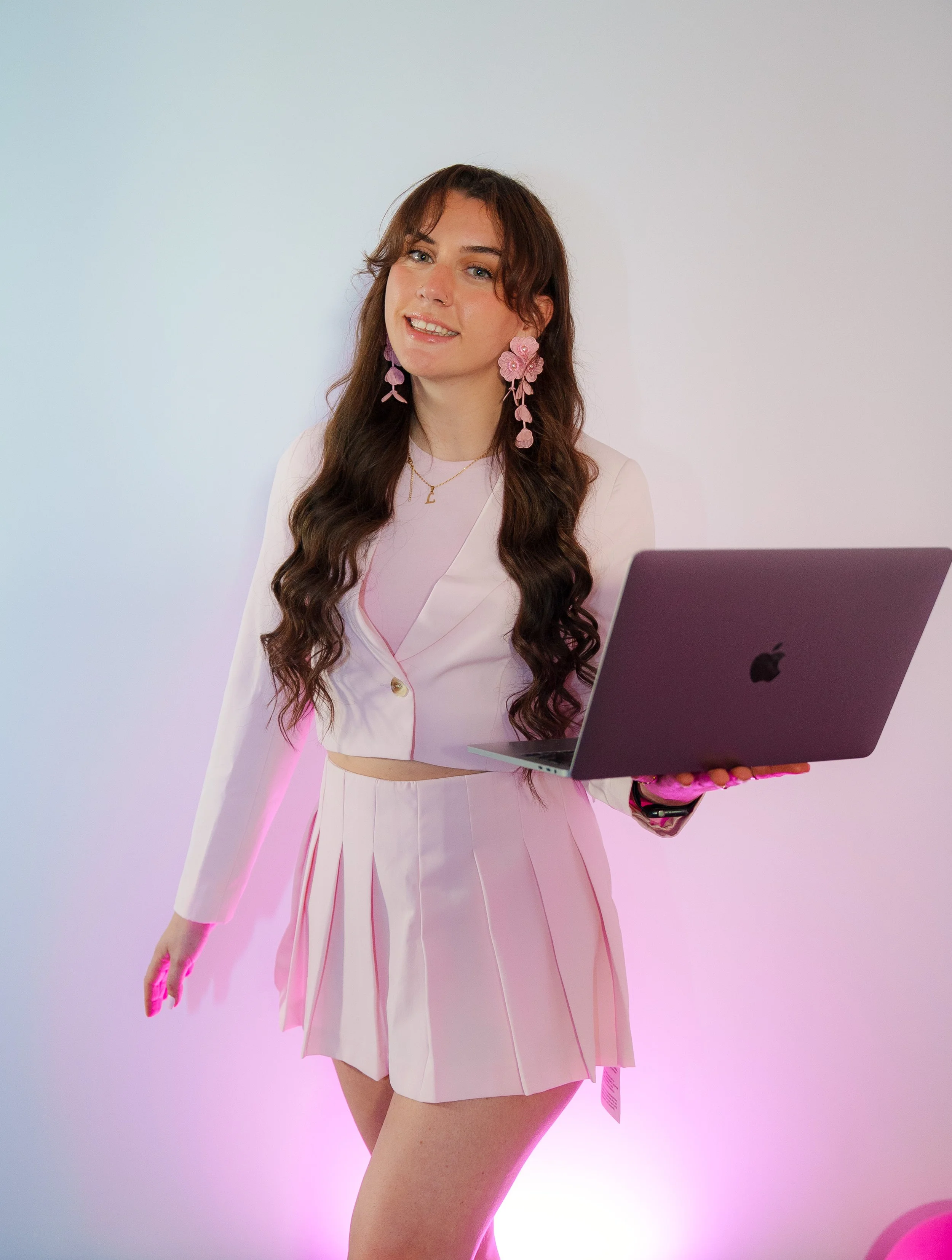 Lily Deal wearing pink floral earrings and a pink and white outfit holding a silver laptop, standing against a white wall with pink and purple lighting.