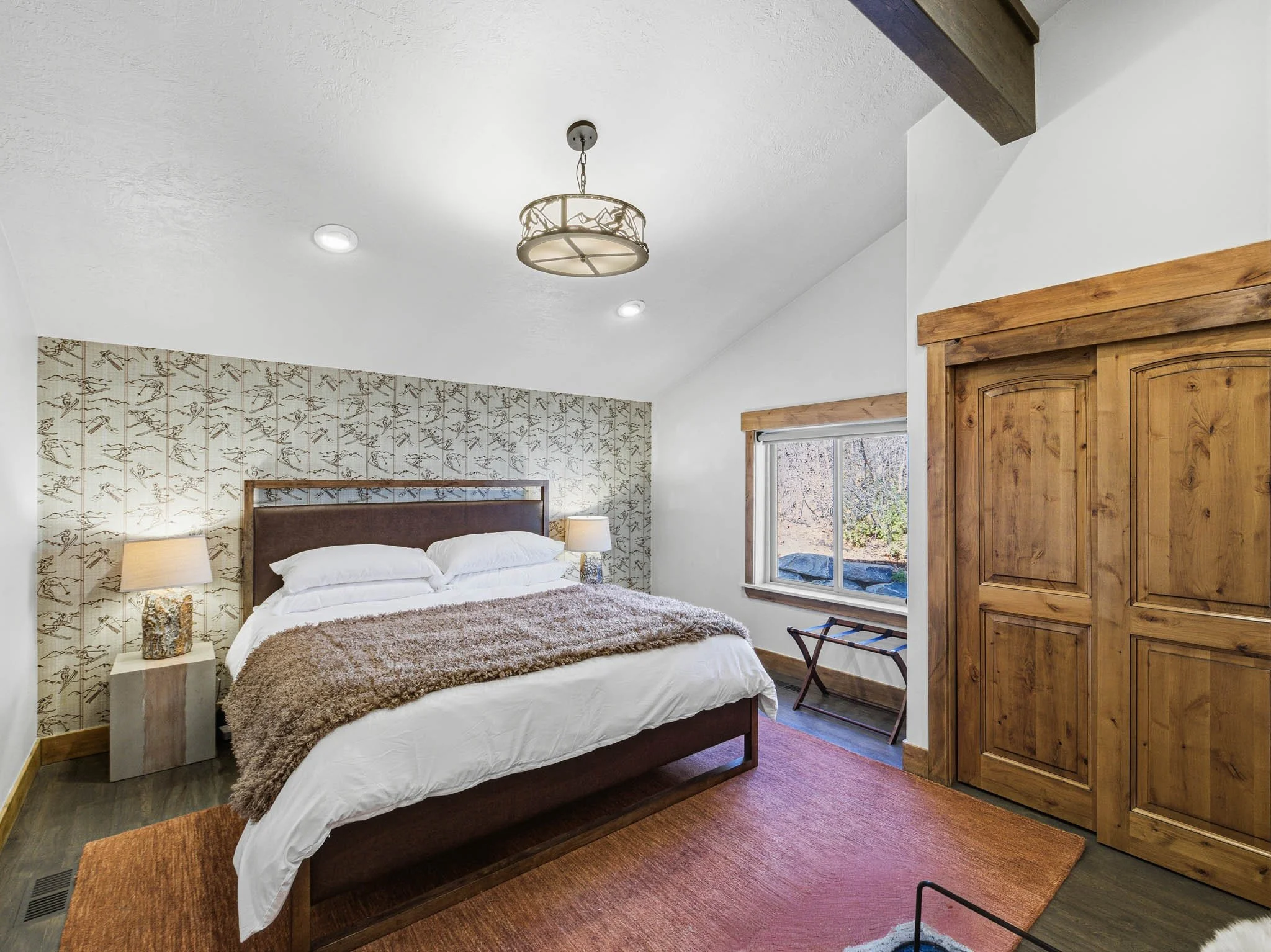 A bedroom with a wood-framed bed, white bedding, and a furry brown blanket, two bedside lamps, a window with a view outside, wood flooring, a large wooden wardrobe, and a ceiling light fixture.