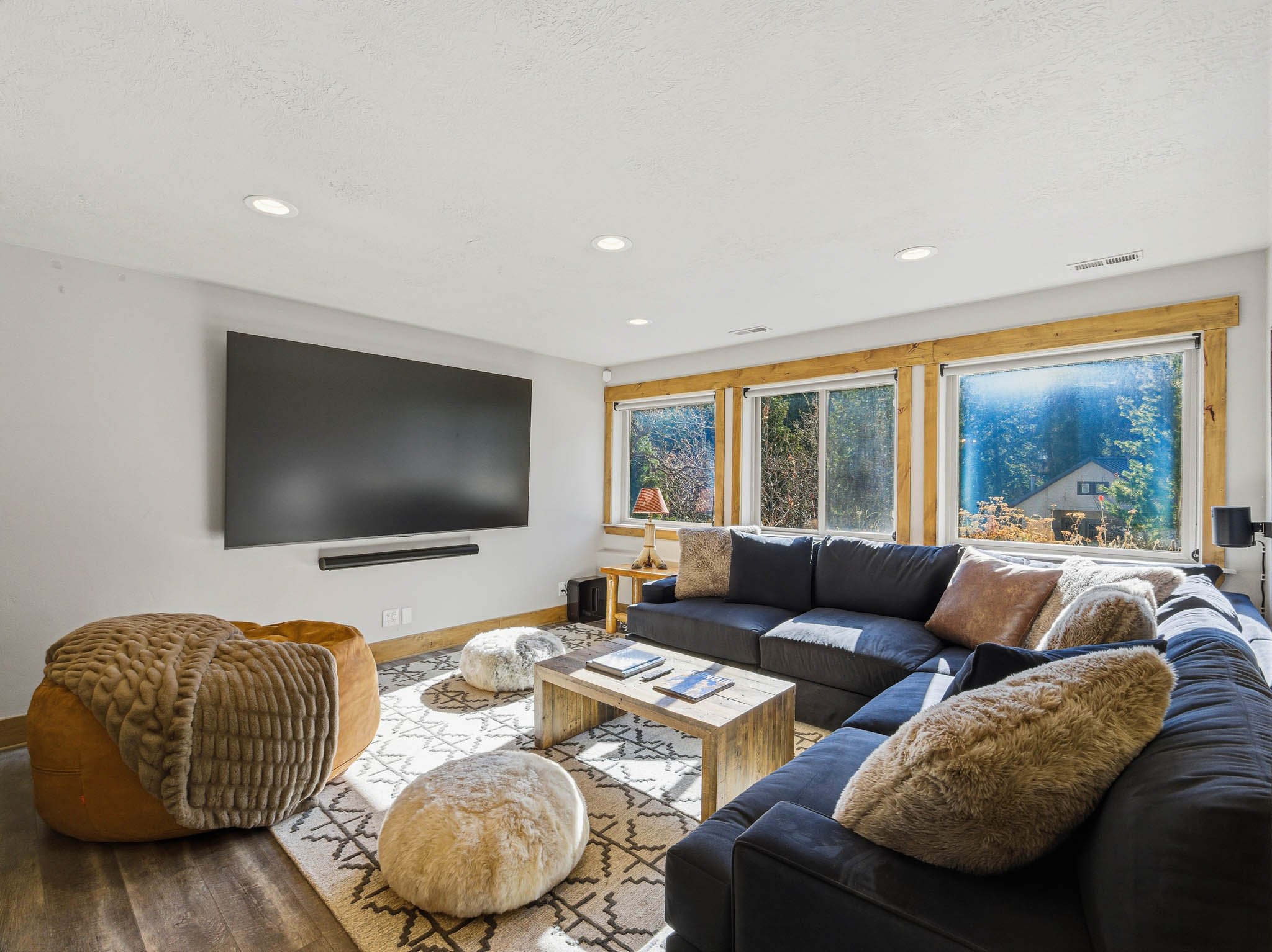 Cozy living room with a large black sectional sofa, plush pillows, fur ottomans, a rustic wooden coffee table, and a wall-mounted flat-screen TV. Large windows with wooden trim let in natural light, showing an outdoor view.