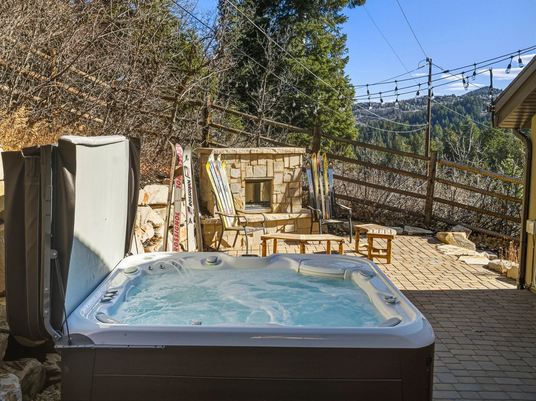 Outdoor patio with a hot tub, stacked skis, small wooden benches, and a stone fireplace, surrounded by a wooden fence and mountain landscape.