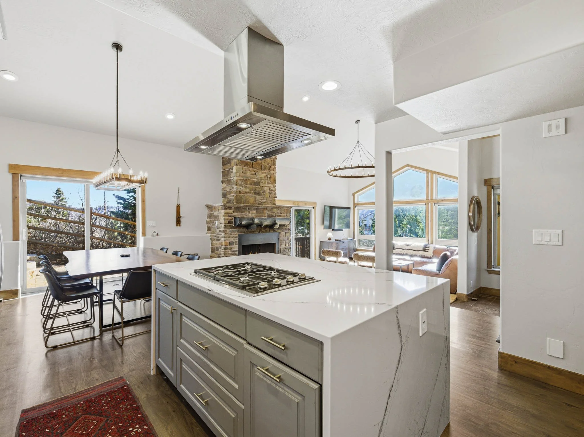 Open-concept kitchen with white island, gas stove, wooden cabinets, stone fireplace, and a bright living area with large windows.