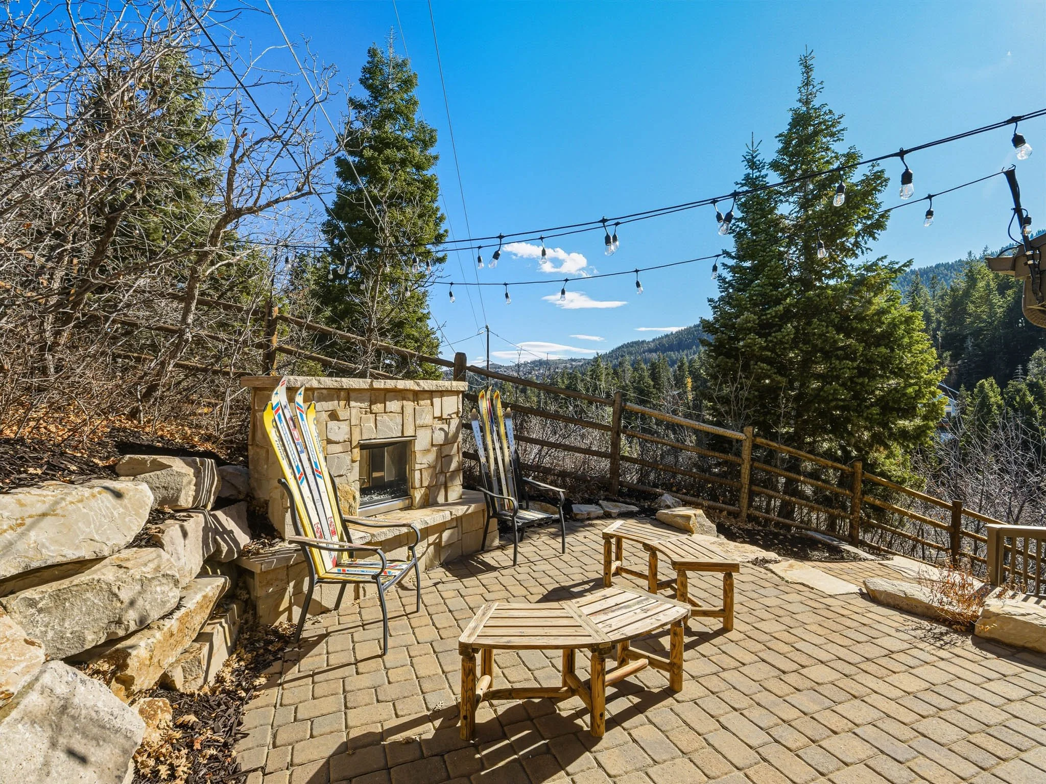 Outdoor patio with pavers, a stone fireplace, two chairs with skis, and a small wooden table, surrounded by trees and mountains, under a string of outdoor lights on a clear day.