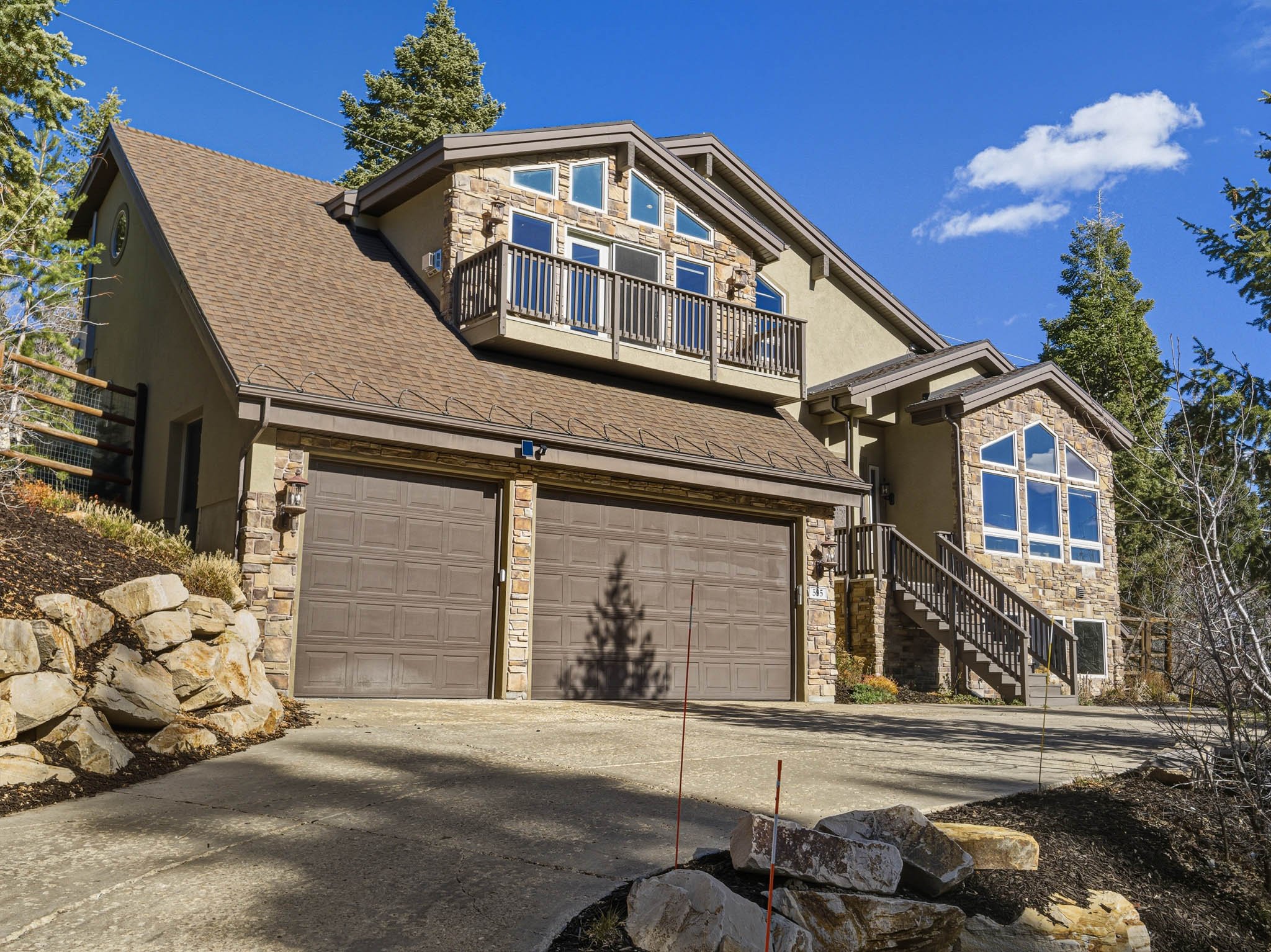 A multi-story house with a stone and stucco exterior, large windows, and a concrete driveway leading to an attached double garage. The house has a small balcony, stairs, and is surrounded by trees and landscaping.