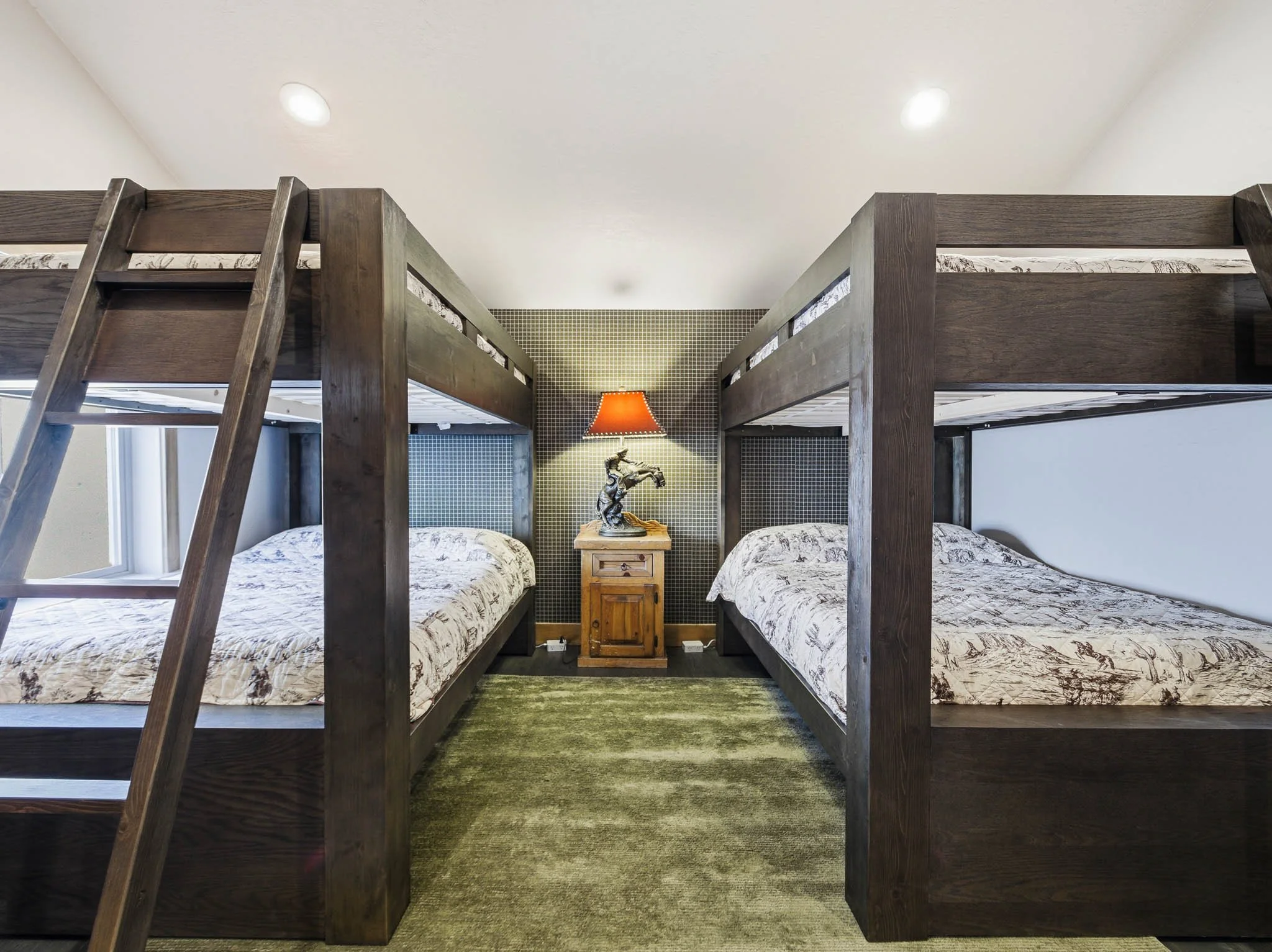 Room with two sets of dark wooden bunk beds, a small wooden nightstand with a lamp and a decorative sculpture, green carpet, white walls, and ceiling with recessed lighting.