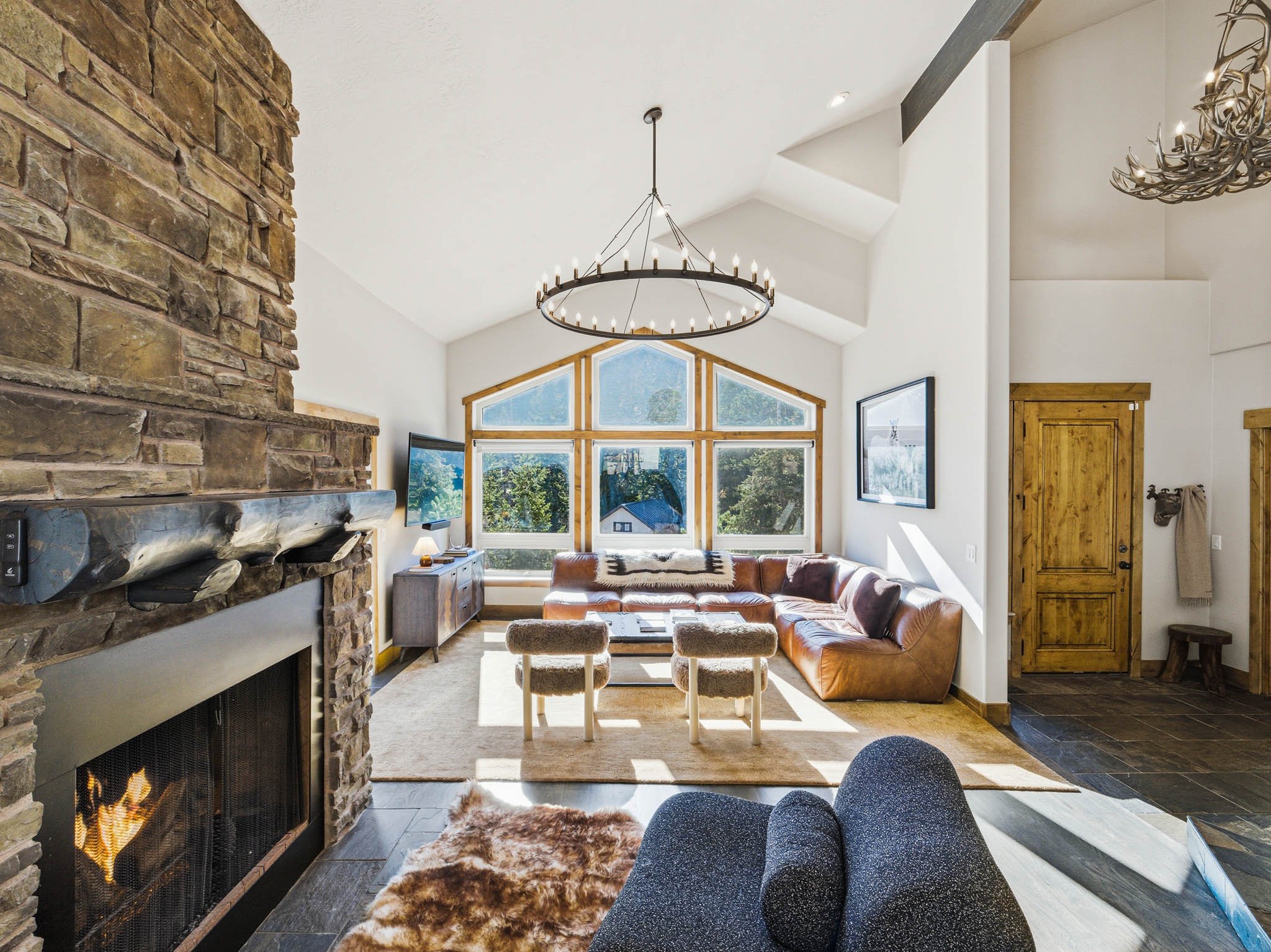 Interior of a bright living room with a stone fireplace, leather sofa, and large window with mountain view.