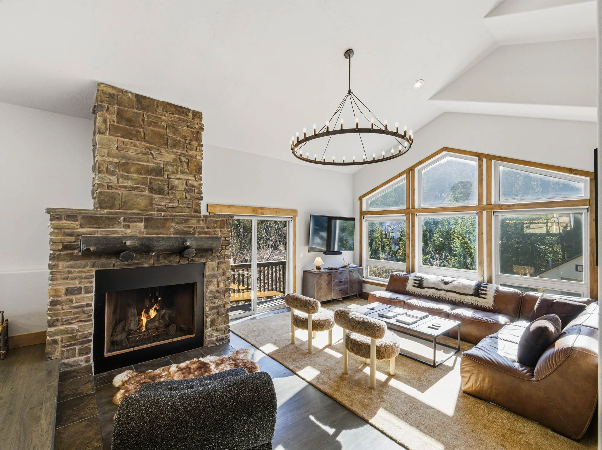 Living room with stone fireplace, leather sofa, wood accents, large windows, and modern light fixture.