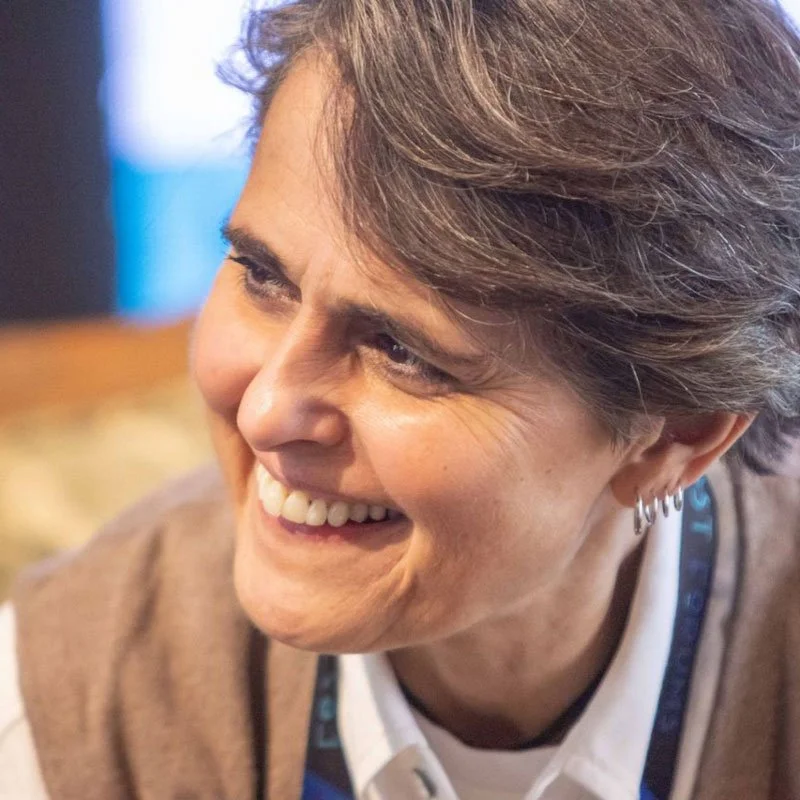 Close-up of a woman smiling with short, gray hair, wearing earrings and a beige and white outfit.
