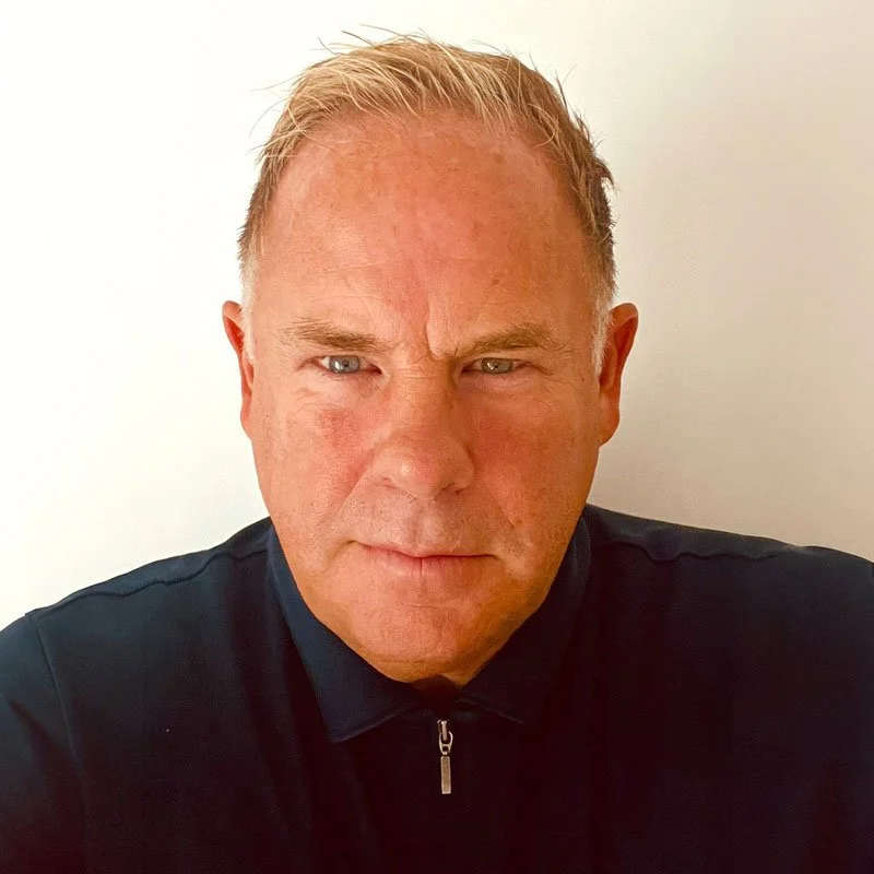 A close-up portrait of a middle-aged man with short, light-colored hair, wearing a black zip-up top, against a plain white background.