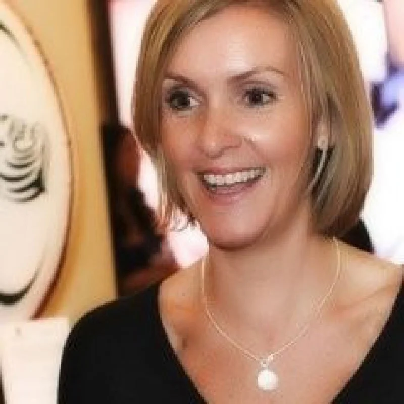 A woman with shoulder-length blonde hair smiling, wearing a black top and pearl necklace, standing indoors with people and artwork in the background.