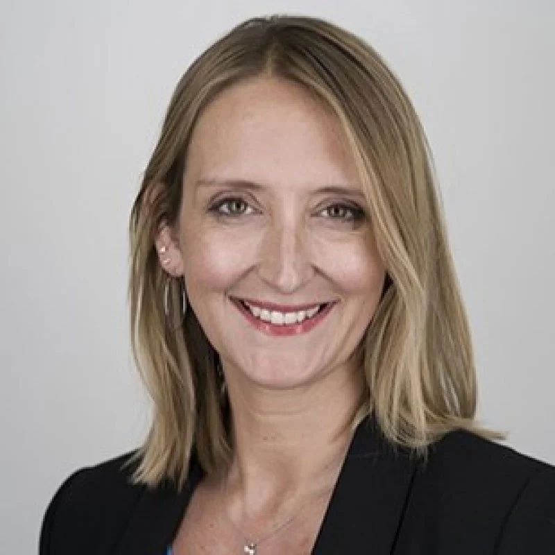 Portrait of a woman with blonde hair, smiling, wearing a black blazer and a necklace, against a plain white background.