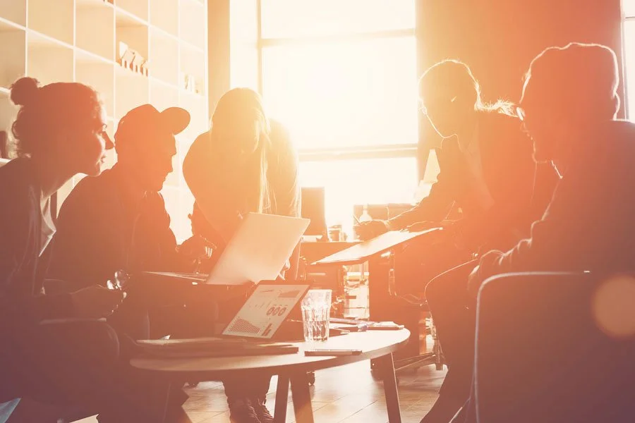 Group of five people in a sunlit room working together around a table with laptops and documents.