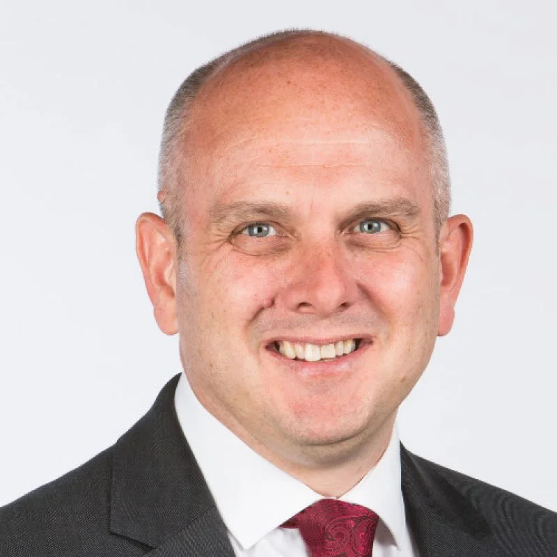 A professional headshot of a middle-aged man with short, light-colored hair and blue eyes, wearing a dark suit, white shirt, and red tie, smiling against a plain white background.