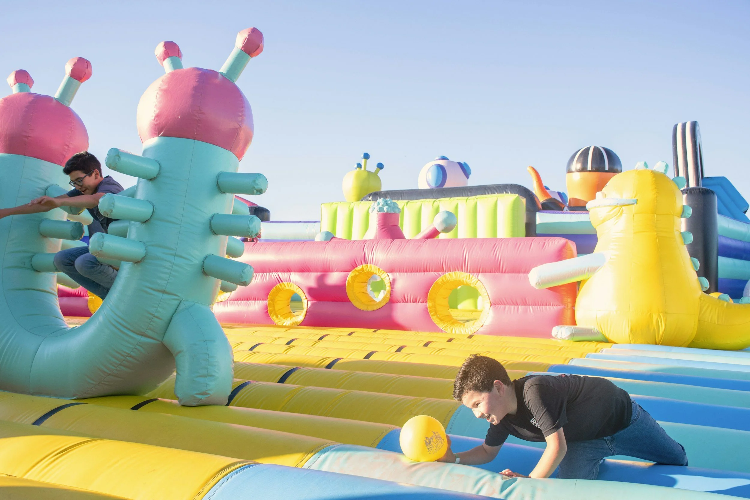 Children playing on a colorful inflatable bounce house designed as a whimsical castle with large pastel pink, blue, yellow, and green elements under a clear sky.