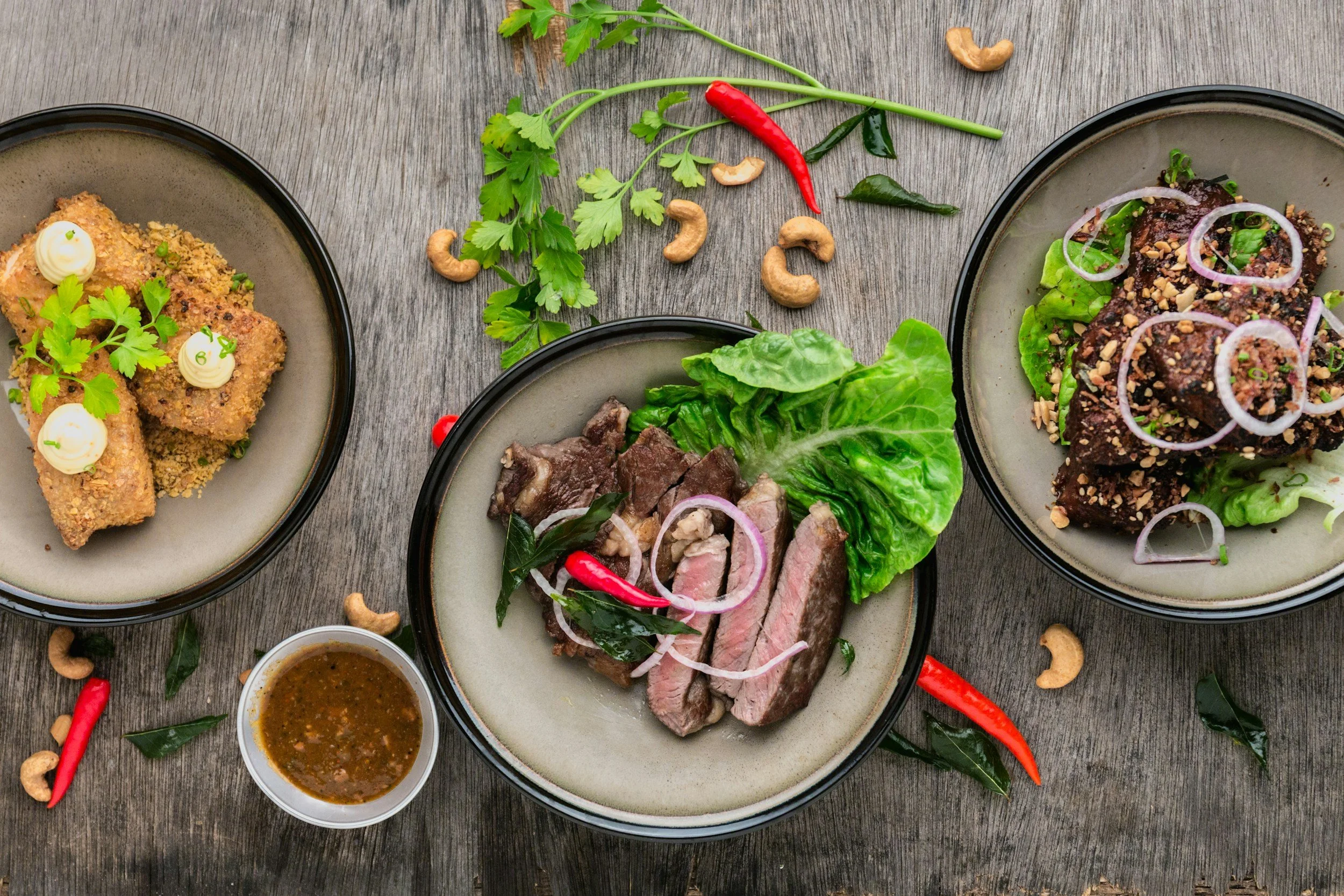 Three bowls of Asian dishes on a wooden table with chili peppers, cashew nuts, fresh herbs, and sauce scattered around.