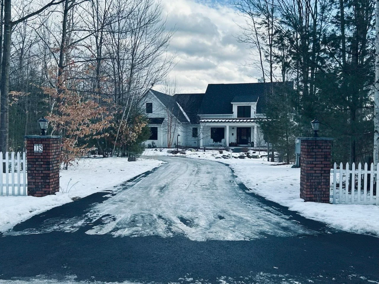 A snow-covered driveway leading to a large white house with a black roof, surrounded by trees and brick pillars with lanterns at the entrance.