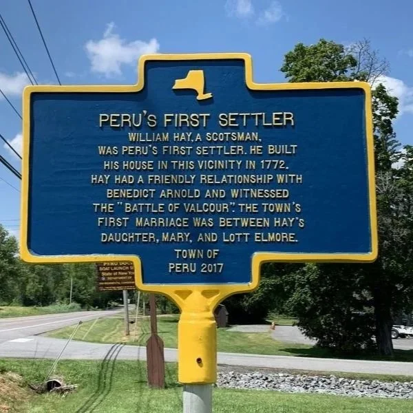 Blue historical marker with yellow border about Peru's first settler, William Hay, built his house in 1772, and his relationship with Benedict Arnold, located on a roadside with trees and other signs in the background.
