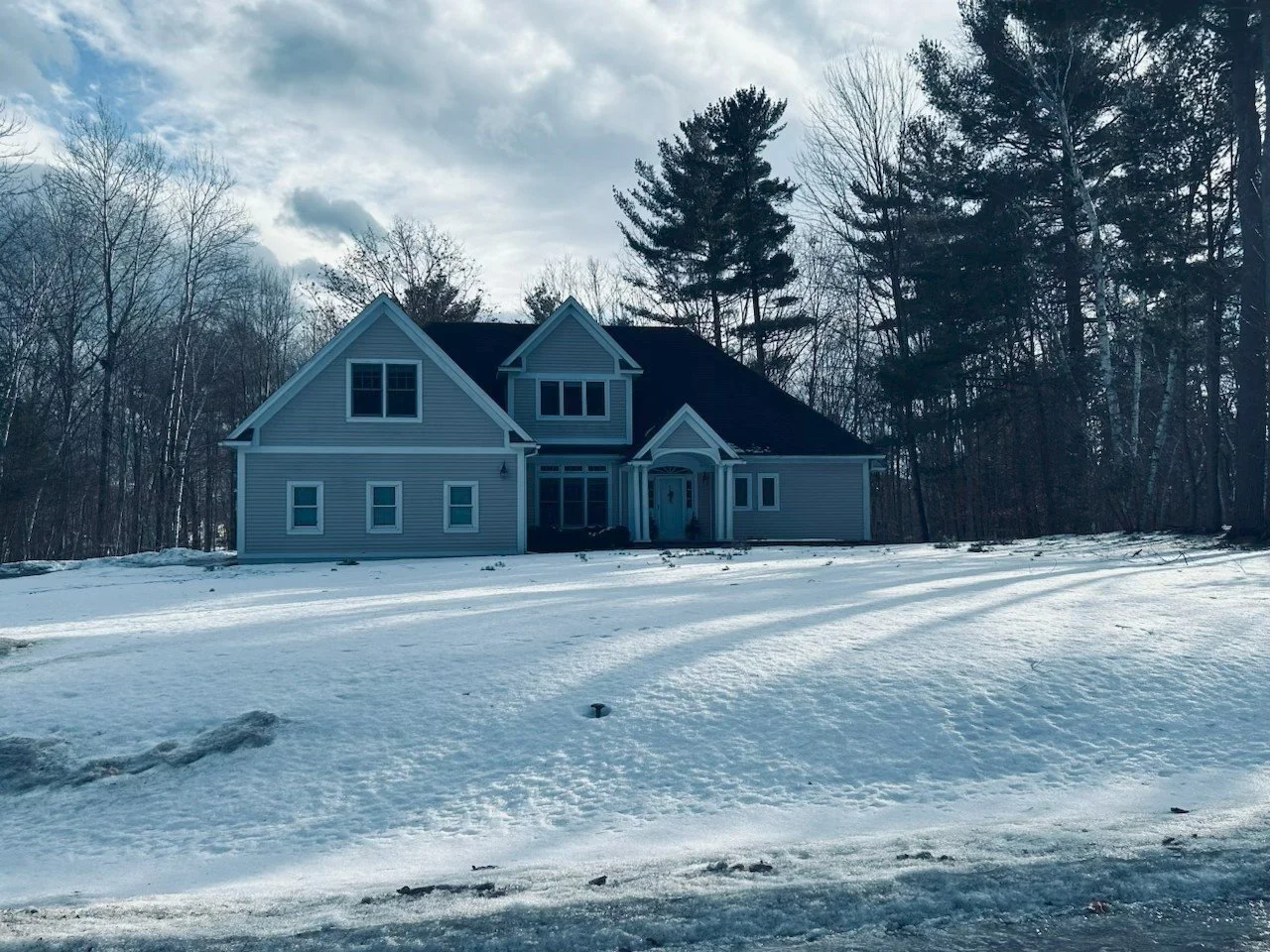 A large house with grey siding and a black roof surrounded by a snow-covered yard and leafless trees, under a cloudy sky.