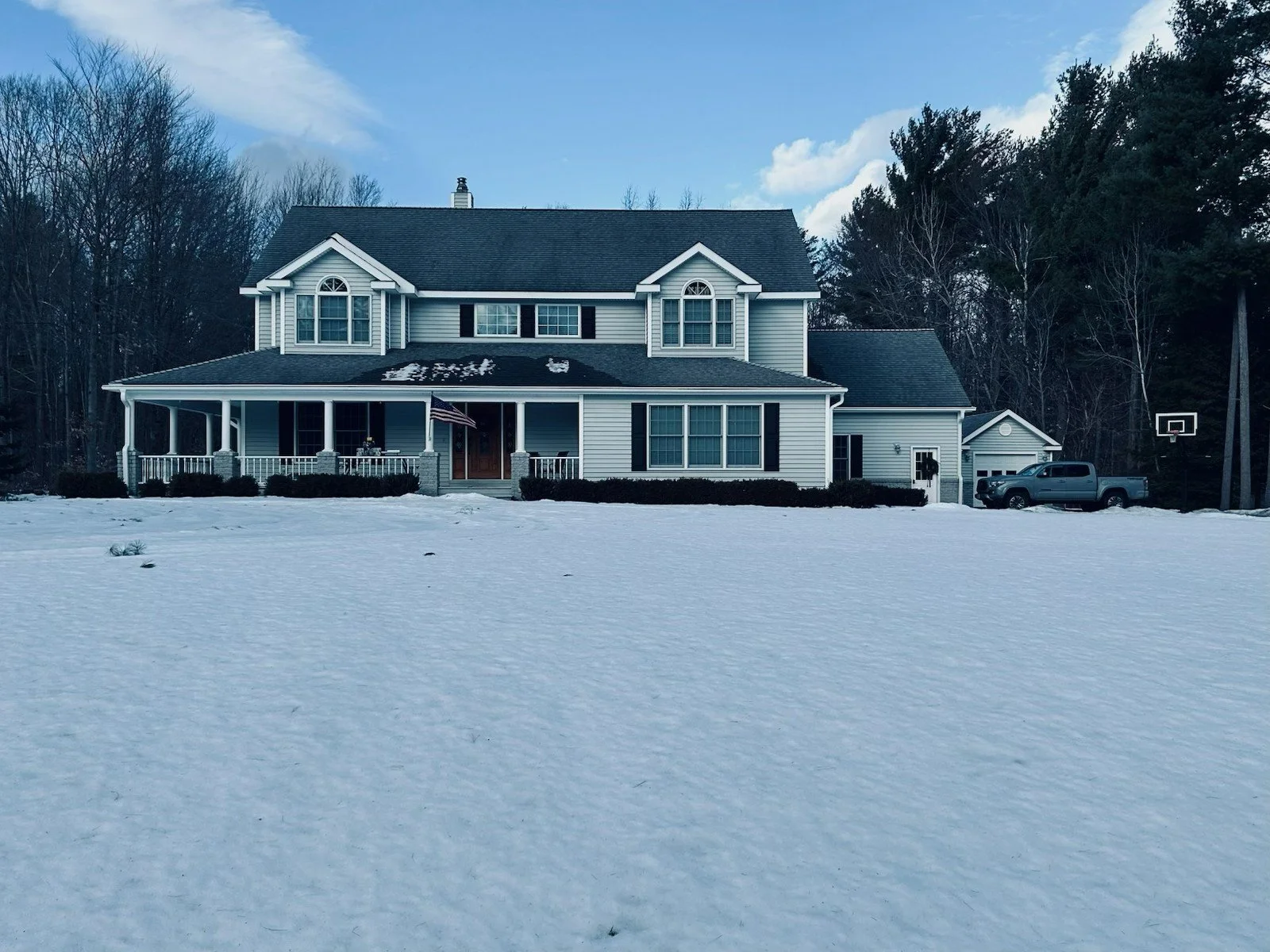 A large two-story house with white siding and black shutters, surrounded by snow, with a front porch and an American flag, a driveway with a truck, and a basketball hoop in the yard.