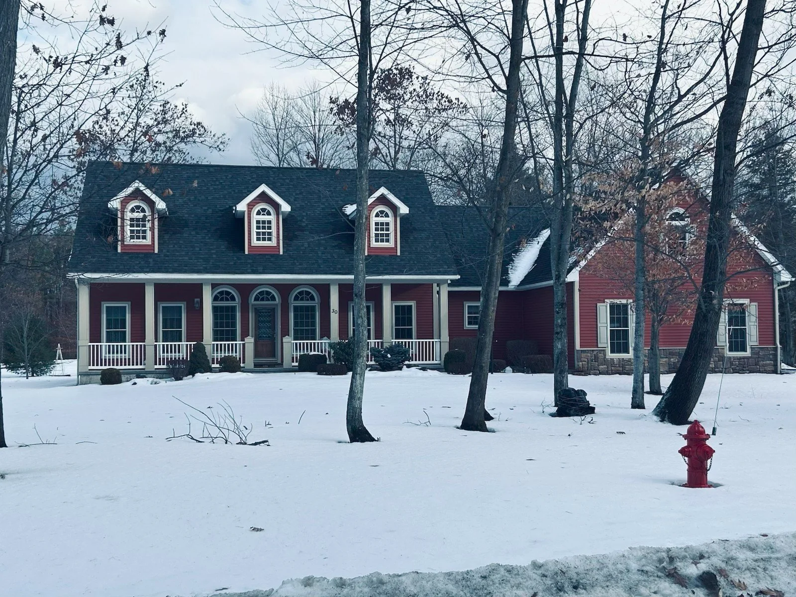 A large red house with white accents and a black roof is surrounded by snow in winter, with leafless trees in the foreground and background, and a red fire hydrant in the front yard.