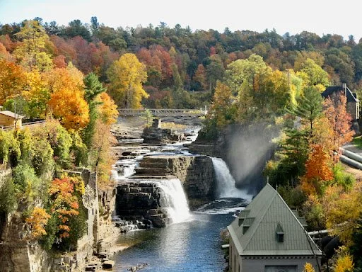A waterfall flowing over rocks in a river surrounded by colorful autumn trees with a green building in the foreground.