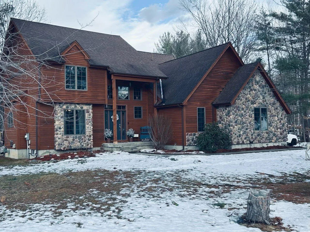 Two-story house with wooden siding and stone accents, snow-covered yard, leafless trees, and a white truck in the background.