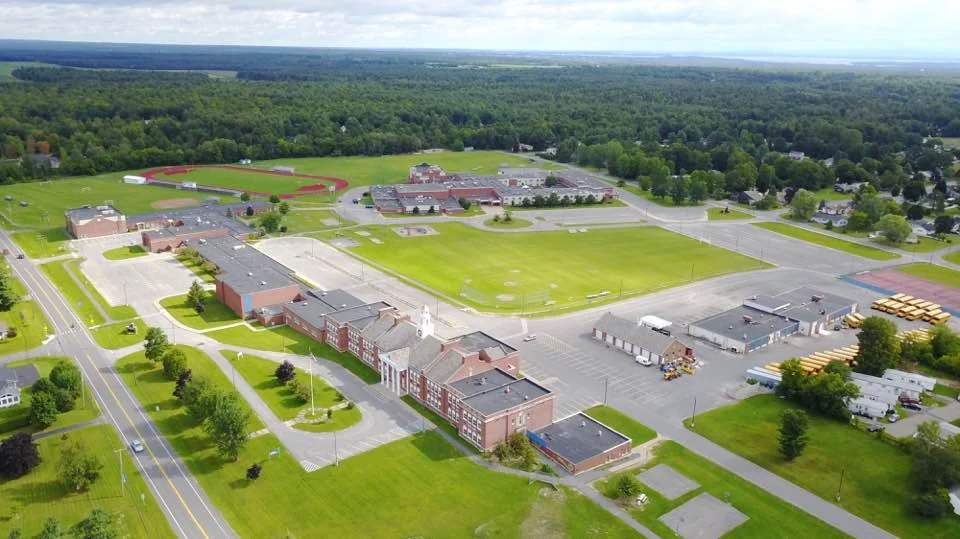 Aerial view of a school campus with a large building, sports field, parking lots, and surrounding greenery.