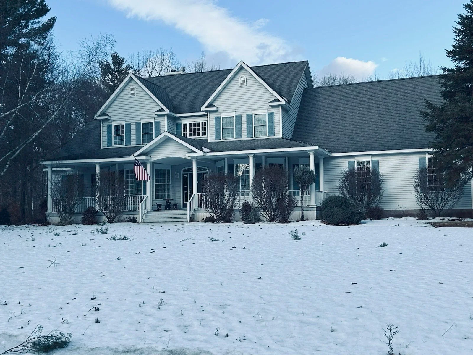 A large two-story house with white siding, blue shutters, and a dark gray roof, surrounded by snow on the ground and leafless trees in the background.