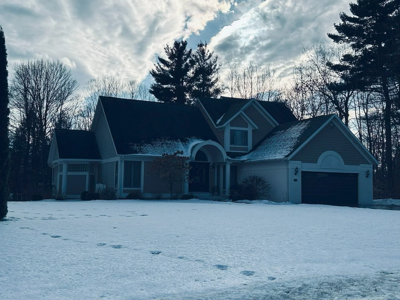 A two-story house with gray siding, a black front door, and a two-car garage, surrounded by leafless trees and snow on the ground and roof, with footprints leading up to the house, under a partly cloudy sky.