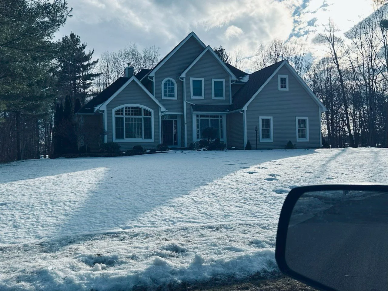 A large, multi-story house with blue siding and white trim is shown from the road with a snowy lawn in the foreground. The house has multiple windows, including a large arched window, and a steep roof. A side mirror of a vehicle is visible in the bot